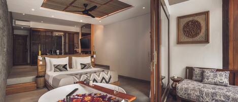 A modern bedroom with a large bed, a sitting area with a sofa, a bathtub with flowers, and a ceiling fan.