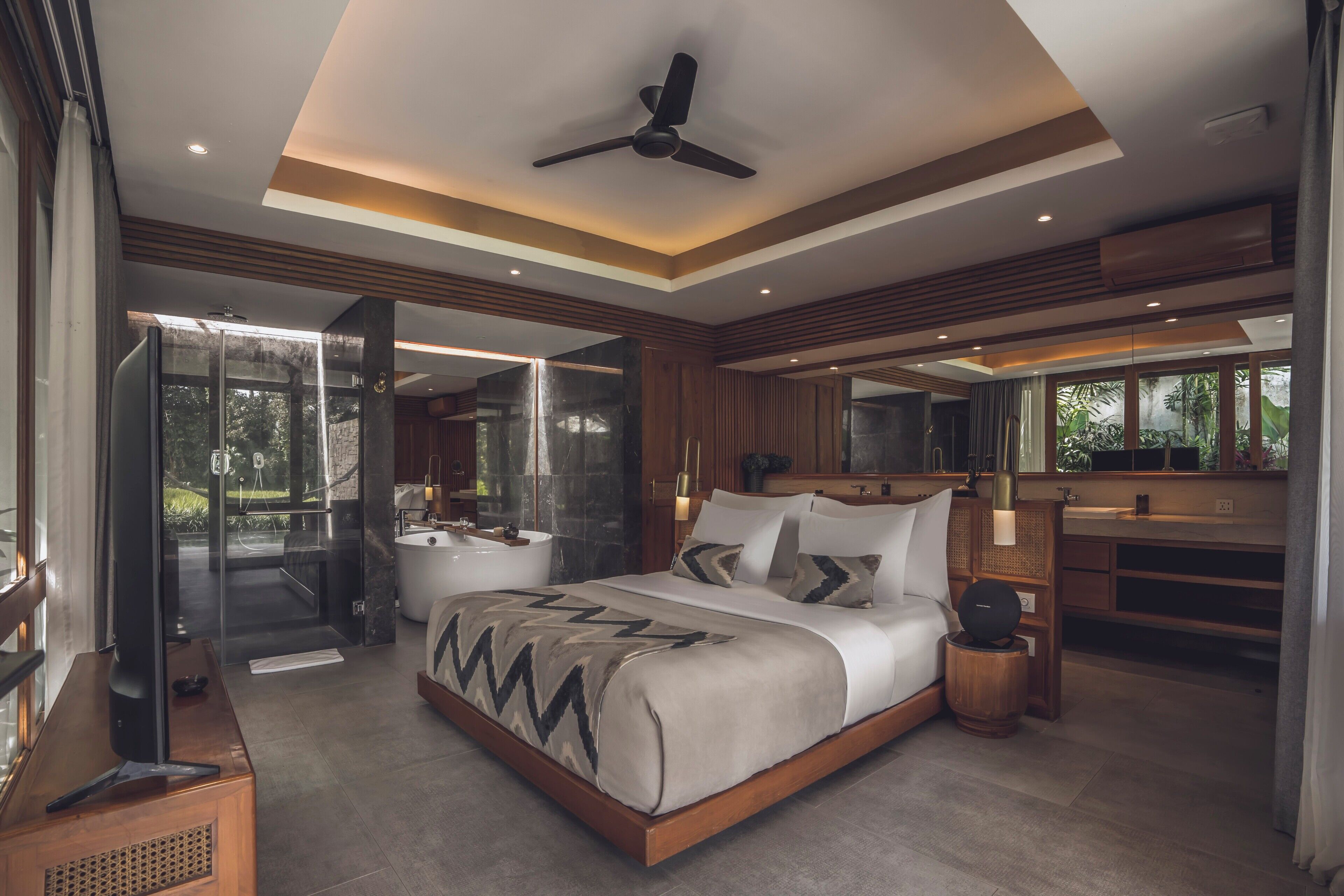 Un dormitorio moderno con una cama grande, un ventilador de techo, cabina de ducha de vidrio y baño con un espejo grande y lavamanos.