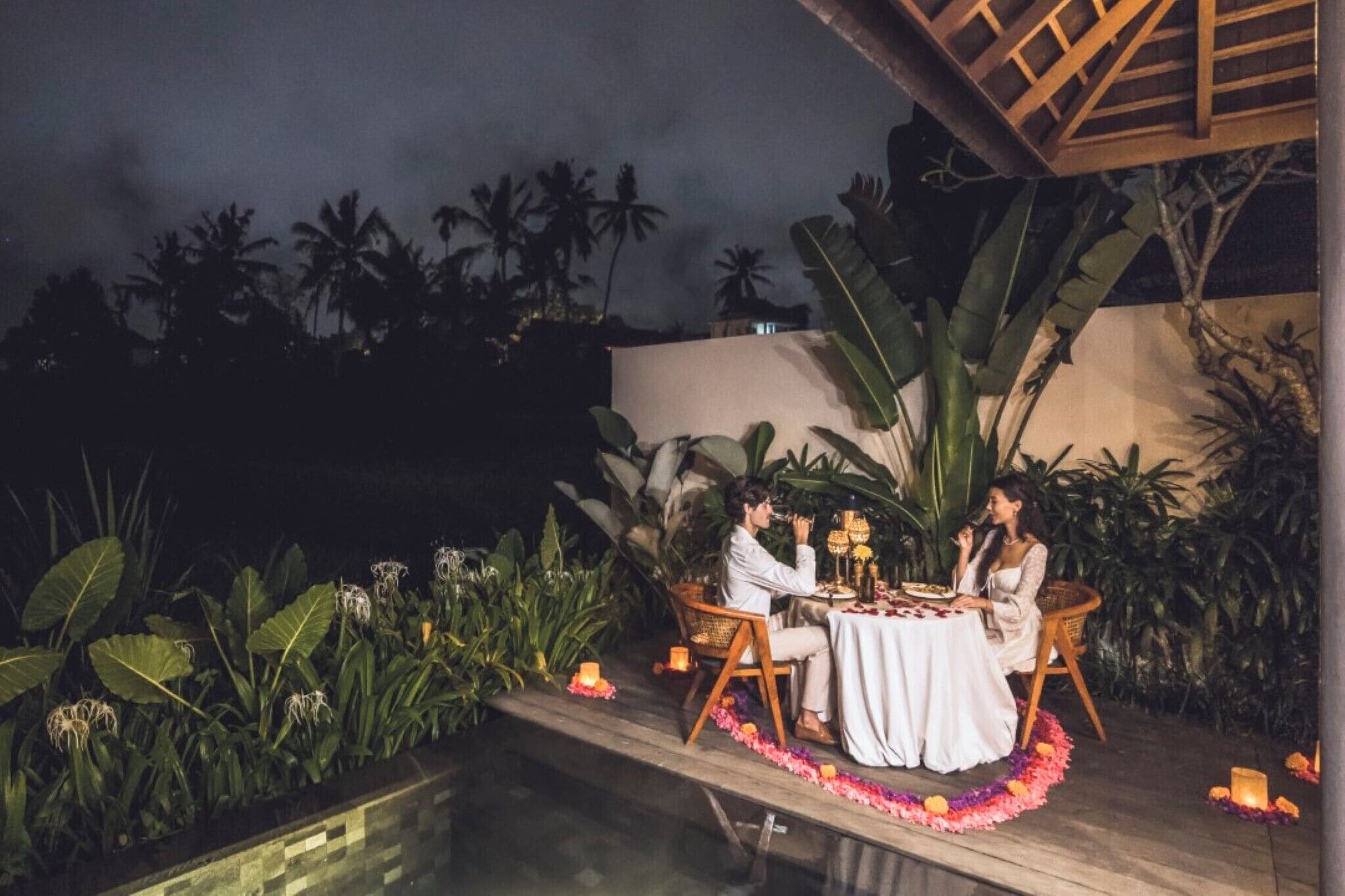 An outdoor dining area with a table set for two, surrounded by tropical plants and a wooden deck.