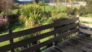 Un banc en bois donnant sur un jardin luxuriant, où poussent de nombreuses espèces de plantes, et d’où l’on aperçoit au loin des maisons et des montagnes.