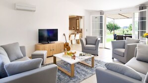 Una sala de estar moderna con un televisor de pantalla plana, sofás grises, una mesa de centro con frutas y revistas, una guitarra y un comedor visible a través de una puerta de vidrio.