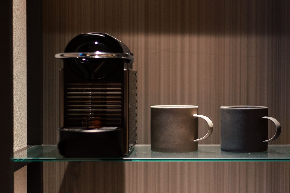 A coffee maker on a glass shelf with two mugs beside it.