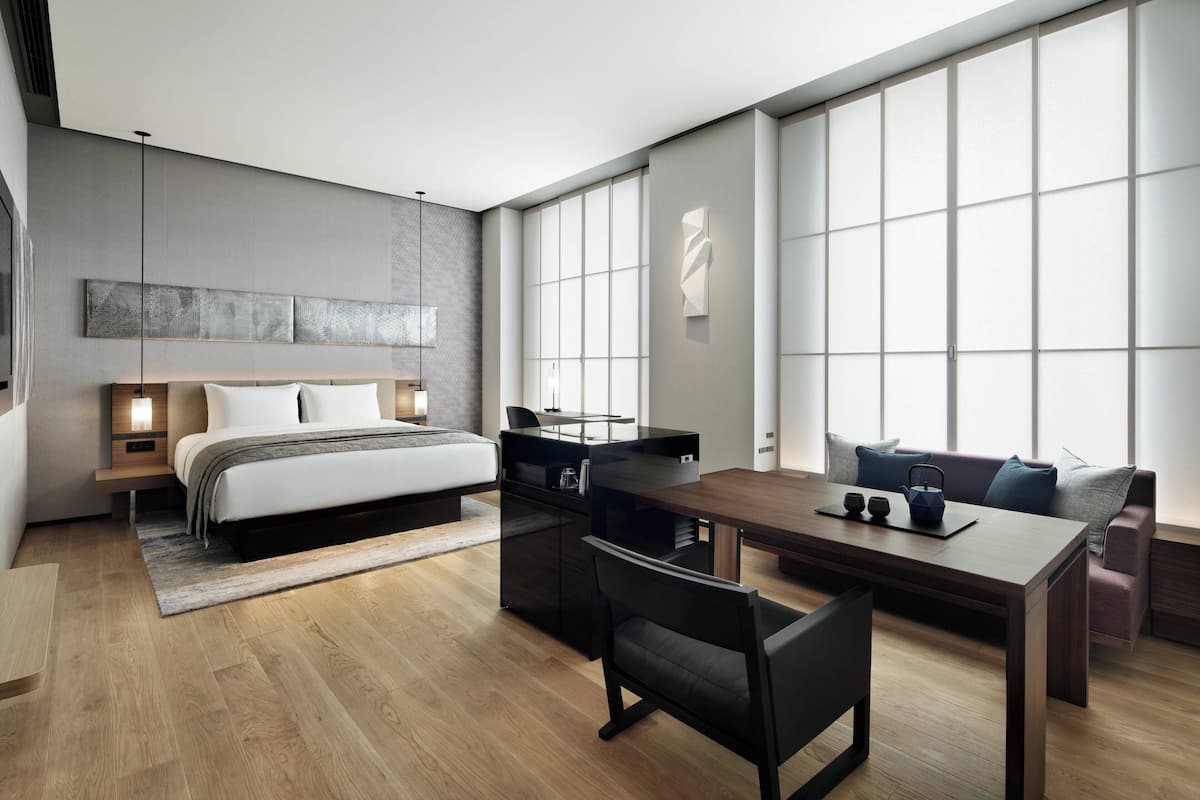 Een moderne hotelkamer met een groot bed, een bureau met stoel, een bank en een groot raam dat natuurlijk licht binnenlaat.