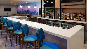 Un espace bar avec un long comptoir, des chaises bleues, un téléviseur et des étagères remplies de bouteilles.