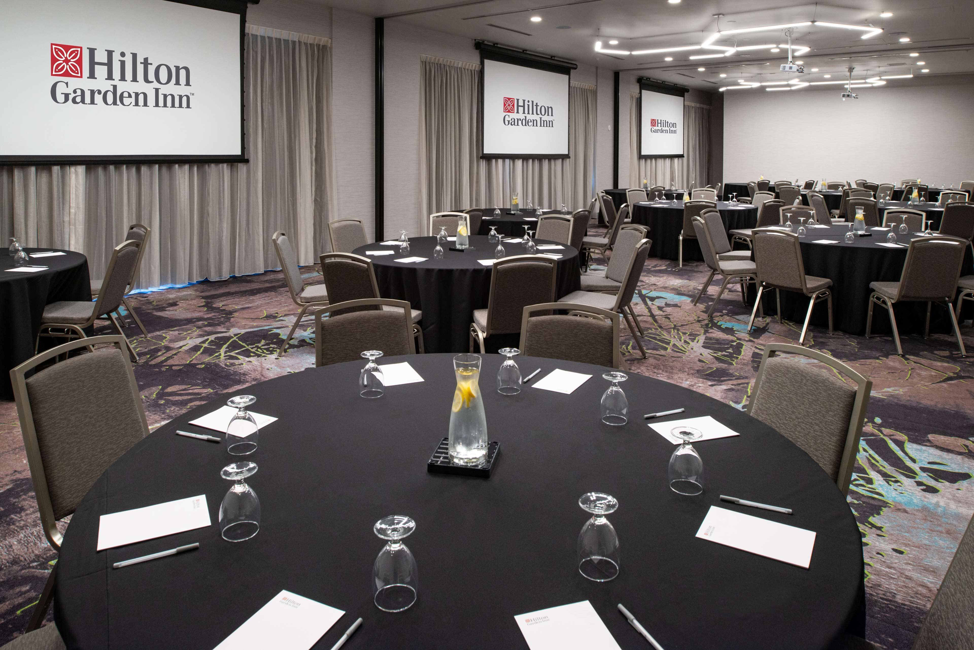 Un salón de conferencias en Hilton Garden Inn con varias mesas, sillas y pantallas para presentaciones.