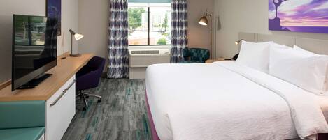 A hotel room with a bed, desk, TV, and a window with curtains.