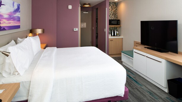Quarto de hotel com uma cama grande, televisão, secretária e uma área de cozinha compacta.