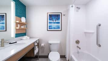 Un baño moderno con lavamanos, inodoro y bañera blancos. Hay estantes con artículos de aseo y un cuadro enmarcado en la pared.