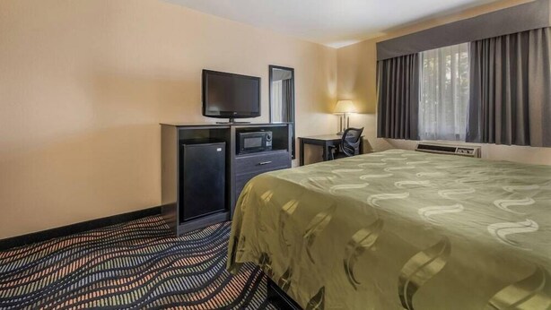A hotel room with a bed, a desk with a chair, a television, a small refrigerator, and a window with curtains. - Rodeway INN Rockford (Rockford)