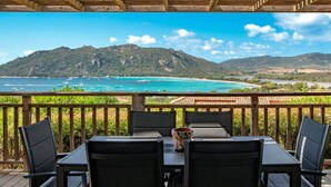 Una terraza de madera con una mesa y sillas que da a un paisaje costero con montañas y aguas azules cristalinas.