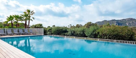 A clear blue swimming pool with a wooden deck, surrounded by palm trees and a white fence, with a mountain in the background.