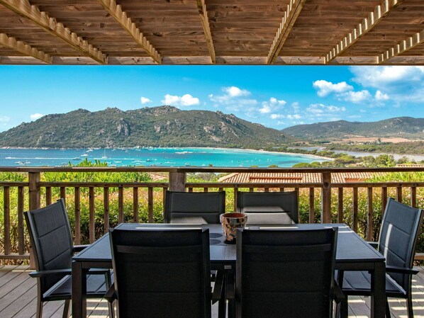 A wooden deck with a table and chairs overlooking a coastal landscape with mountains and clear blue waters.
