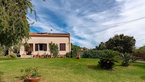 Une maison à deux étages, avec un toit recouvert de tuiles, un petit jardin devant avec des plantes en pot et un ciel dégagé.