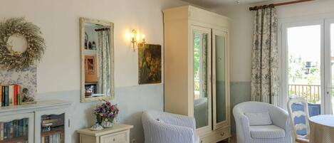 A room with a bed, two striped chairs, a small table, a cabinet with books, a mirror, and a door leading to a balcony.