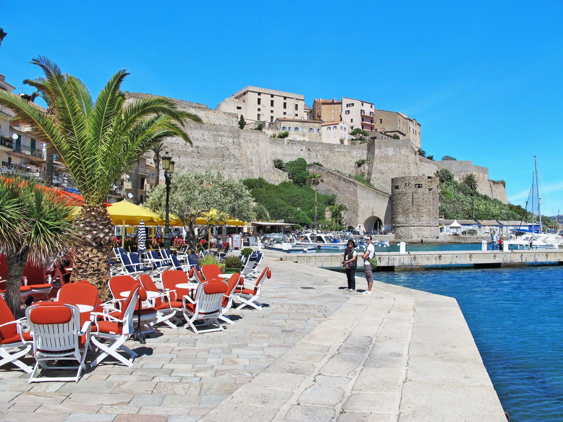 A seaside promenade with outdoor seating, a historic building, and boats in the background.