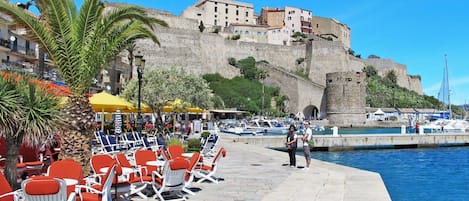 A seaside promenade with outdoor seating, a historic building, and boats in the background.