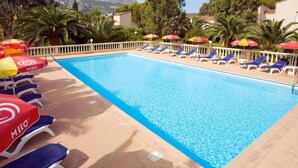 A swimming pool with lounge chairs and umbrellas, surrounded by trees and mountains in the background. - Nice apartment for 2 people with pool, WIFI, A/C, TV and terrace (Calvi)