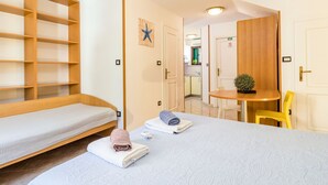 Une chambre d’hôtel avec deux lits simples, une étagère en bois, une petite table avec une plante et une vue sur la cuisine.