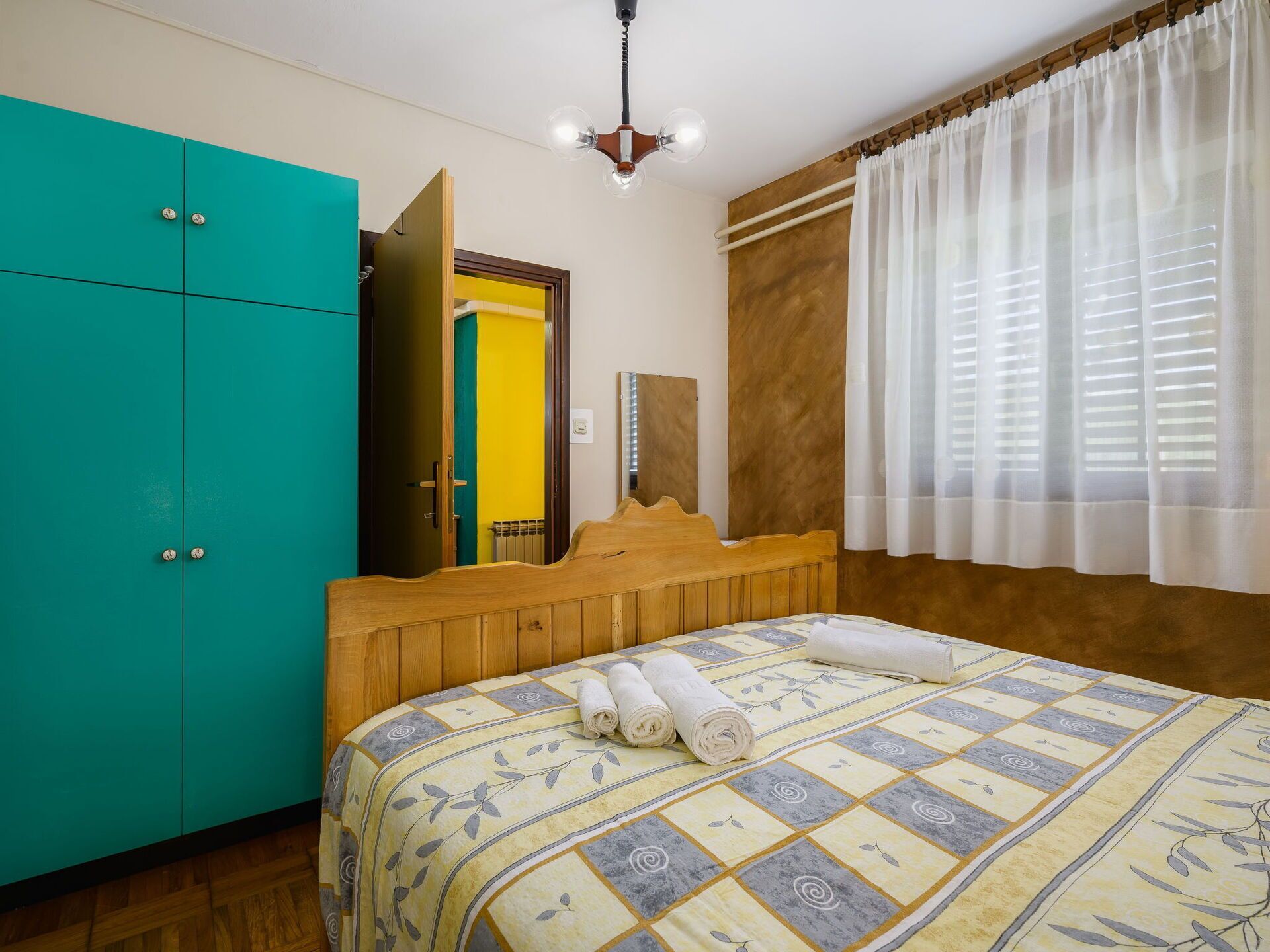 A bedroom with a bed, a teal wardrobe, a wooden headboard, a mirror, and a window with white curtains.