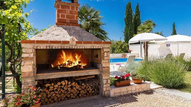 A brick outdoor oven with a fire burning inside, surrounded by a pool area with lounge chairs and umbrellas. - Cosy apartment for 3 guests with pool, A/C, WIFI, TV and terrace (Porec)