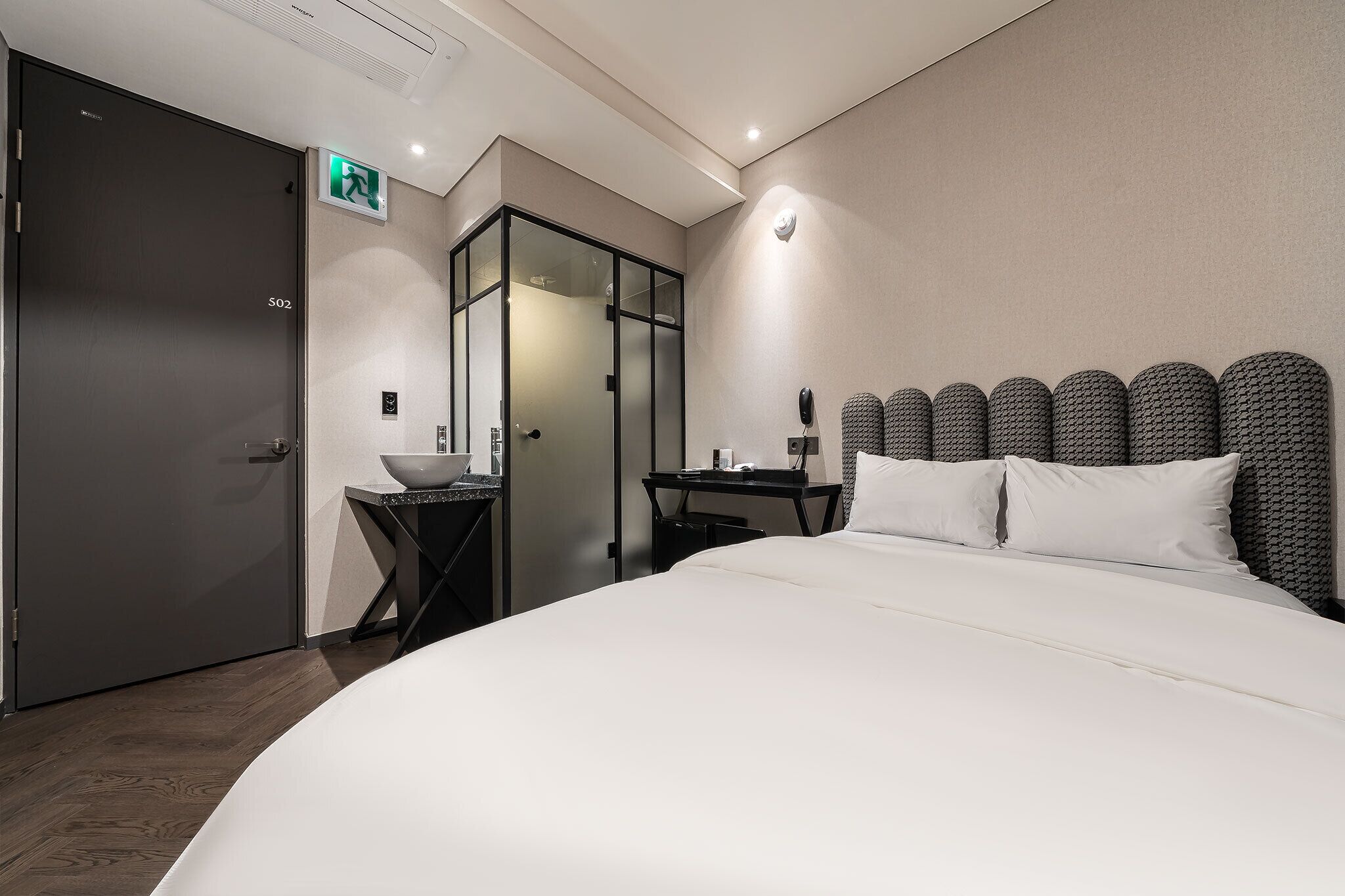 Habitación de hotel con una cama grande, un pequeño escritorio con lámpara, zona de ducha con mampara de vidrio y una puerta identificada con 'S02'.