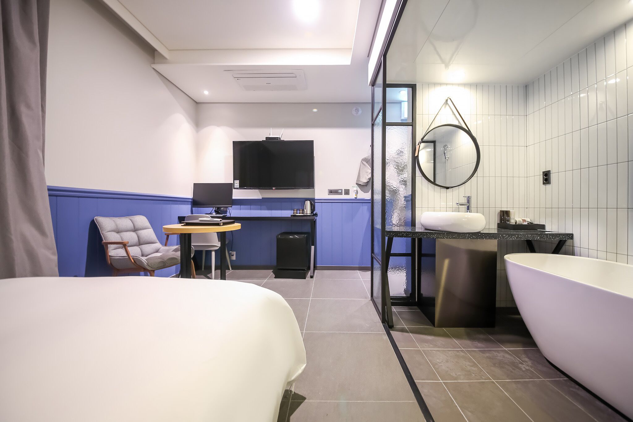 A modern hotel room with a bed, a desk with a flat-screen TV, a small table, a chair, a large bathtub, and a glass-enclosed shower area.