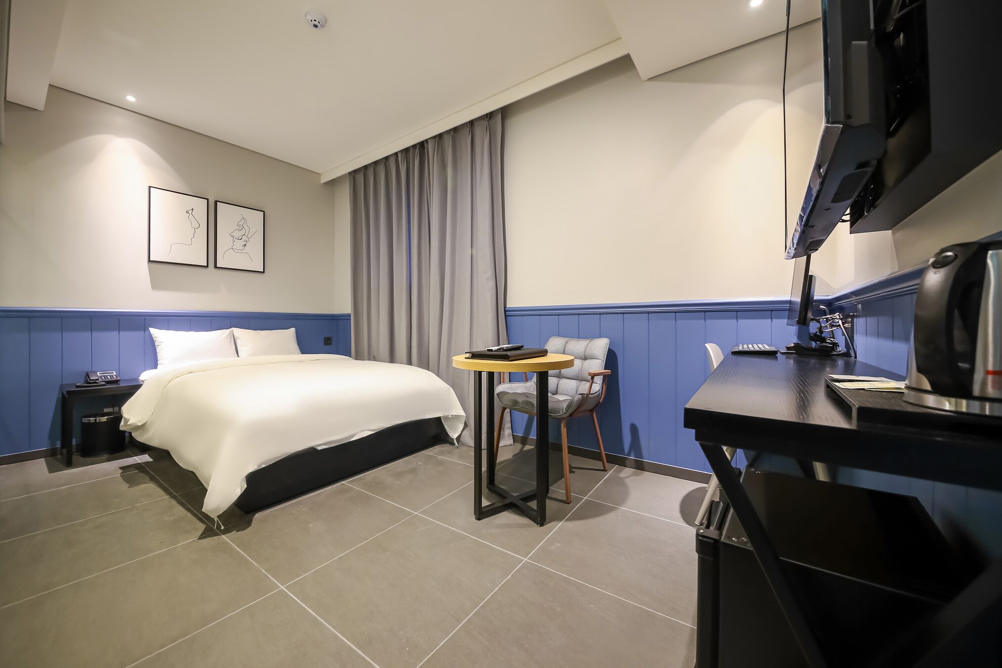 A hotel room with a bed, a small round table, a chair, a desk with a kettle, and a television.
