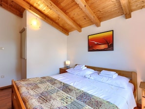 Une chambre avec un plafond en bois, un lit recouvert de draps blancs, une table de chevet avec une lampe, et un tableau représentant un coucher de soleil accroché au mur.