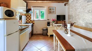 A compact kitchen with a wooden ceiling, a microwave, a stove, and a sink. There's a window with a view of greenery, a television, and a small table with a vase of flowers.