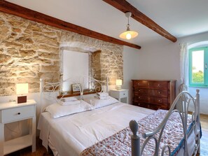 Quarto com parede de pedra, teto de madeira, cama com roupa de cama branca, cômoda e janela com cortinas.