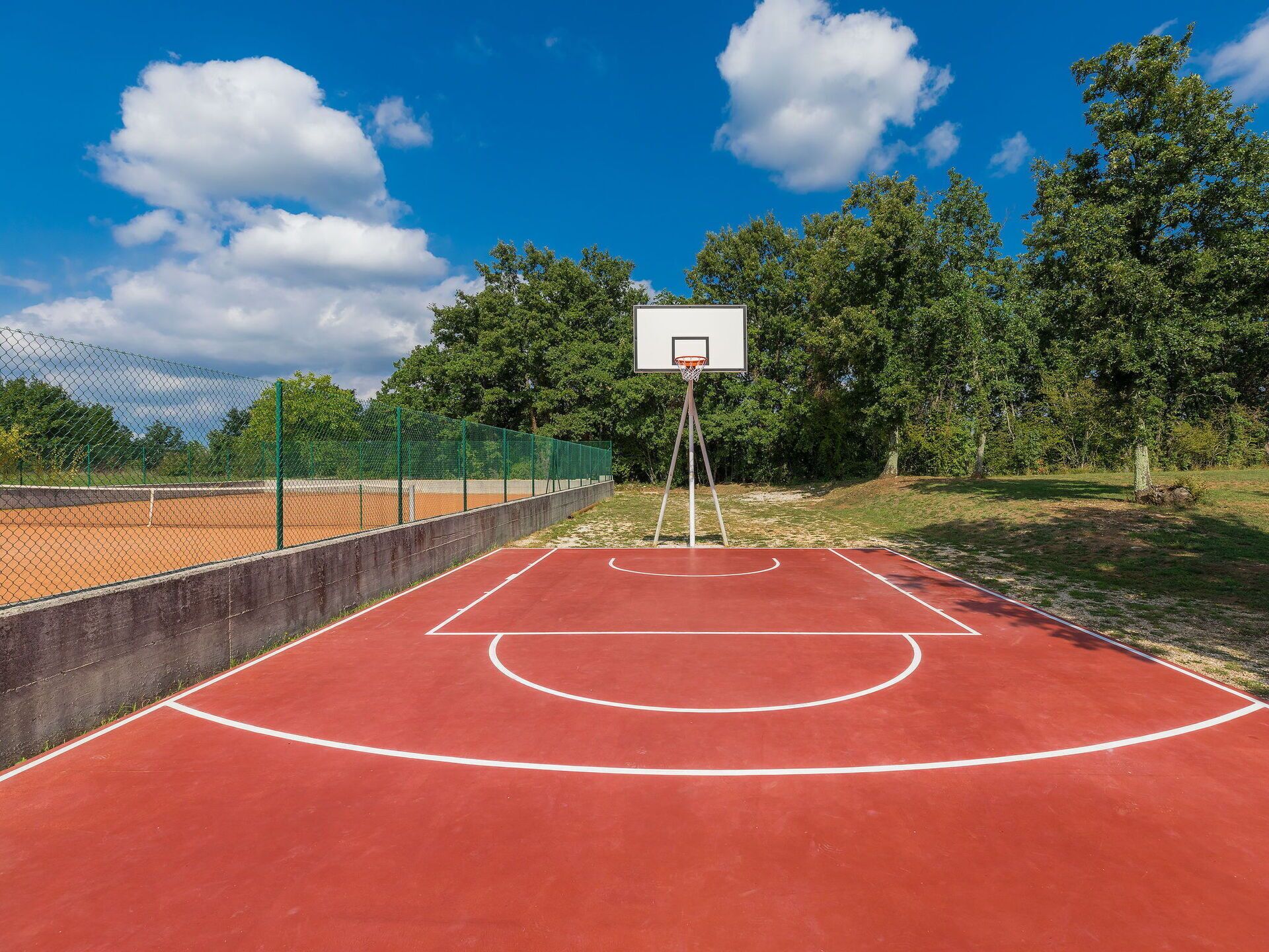 An outdoor basketball court with a red surface, surrounded by a fence and trees.