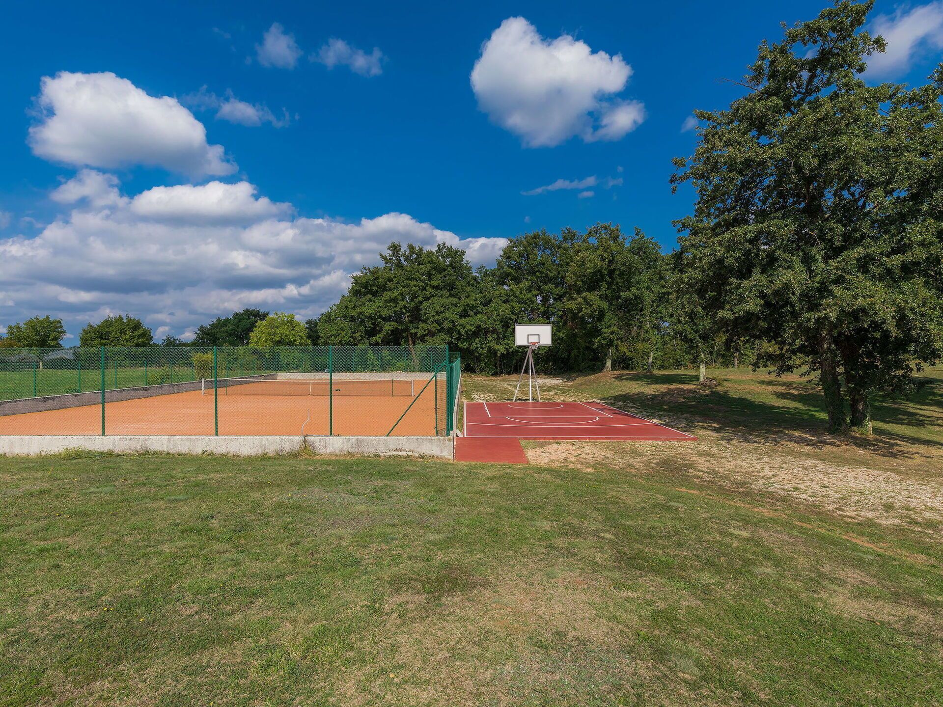 Een tennisbaan buiten met een groene omheining, een basketbalveld en bomen op de achtergrond.