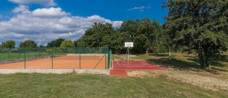 Een tennisbaan buiten met een groene omheining, een basketbalveld en bomen op de achtergrond.
