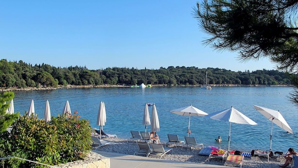 A beachside area with lounge chairs and umbrellas, a stone wall, and a view of the water and trees. - Beautiful apartment for 3 guests with WIFI, A/C, TV, terrace and pets allowed (Rovinj)