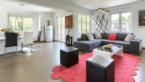 Un salon moderne avec un canapé d’angle gris, une table à manger avec des chaises, une petite table basse noire et un tapis rouge à motifs.