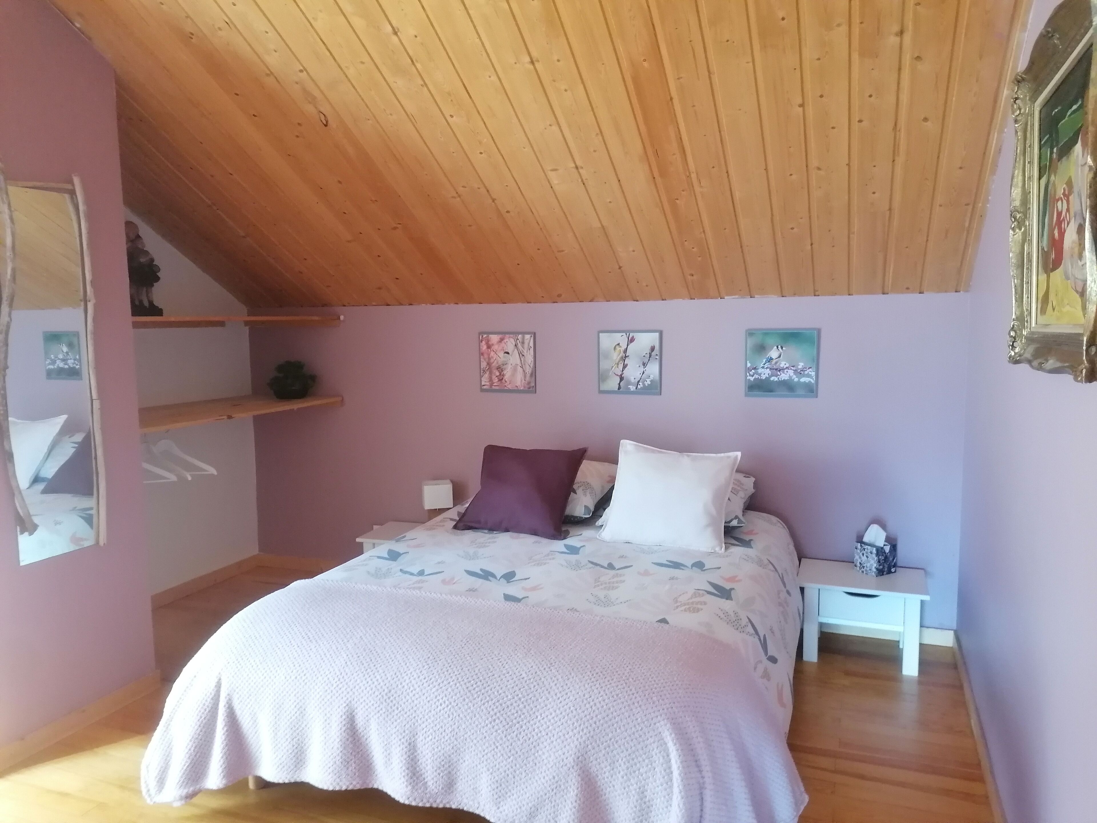 Un dormitorio con techo inclinado, vigas de madera, una cama con ropa de cama floral, una mesita de noche y cuadros en la pared.
