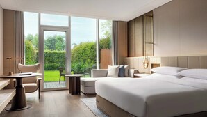 A modern hotel room with a large bed, a desk, a chair, and a view of the garden through the glass door.
