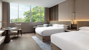 A modern hotel room with two beds, a desk, a chair, and a large window with a view of greenery.