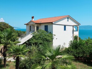 Stunning coastal villa with a scenic view of the sea and distant mountains, surrounded by lush greenery and a prominent staircase leading to the entrance.