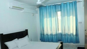 Ein Schlafzimmer mit einem Bett, einem Fenster mit blauen Vorhängen, einem Deckenventilator und einer weißen Wand.