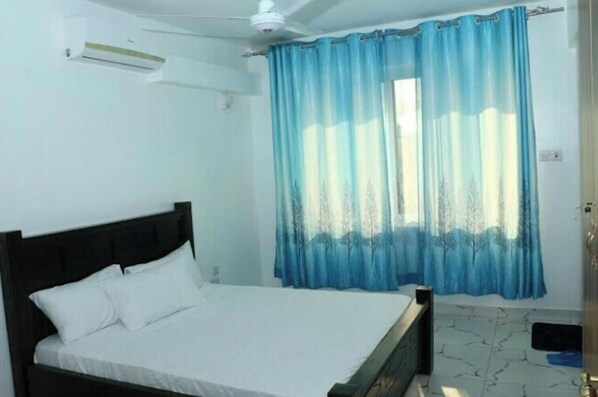 A bedroom with a bed, a window with blue curtains, a ceiling fan, and a white wall. - Hosted   by     Orah (Nyali)