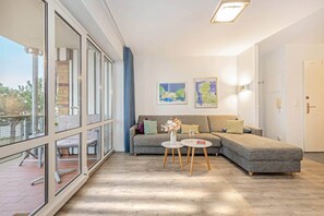 Ein modernes Wohnzimmer mit einem grauen Sofa, einem kleinen runden Tisch mit einer Vase und zwei Gläsern sowie zwei gerahmten Karten an der Wand.