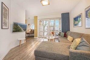 Ein Wohnzimmer mit einer Couch, einem Couchtisch und einem Fernseher. Ein Fenster mit Vorhängen, ein gerahmtes Bild an der Wand und ein kleiner Tisch mit Blumen.