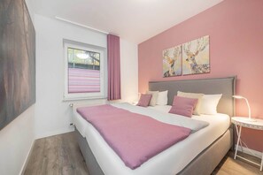 Ein Schlafzimmer mit einem Bett, einem Fenster mit Vorhängen, einem Nachttisch mit Lampe und zwei Wandbildern.