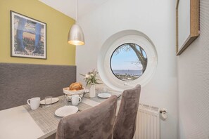 Ein Essbereich mit rundem Fenster, einem Tisch mit Tellern, Gläsern und einer Schüssel Brot, sowie einem gerahmten Bild an der Wand.