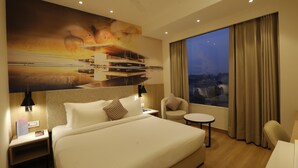 Habitación de hotel con una cama grande, un escritorio, una silla, una mesita y un amplio ventanal con cortinas.