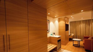 Una habitación de hotel moderna con suelo de madera, un armario de madera, una pequeña kitchenette, una mesa redonda, un sofá y un ventanal con cortinas.