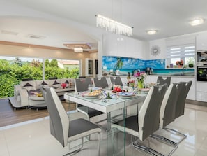 Een moderne eethoek met een glazen tafel en stoelen, een bank en een keuken met uitzicht op groen.