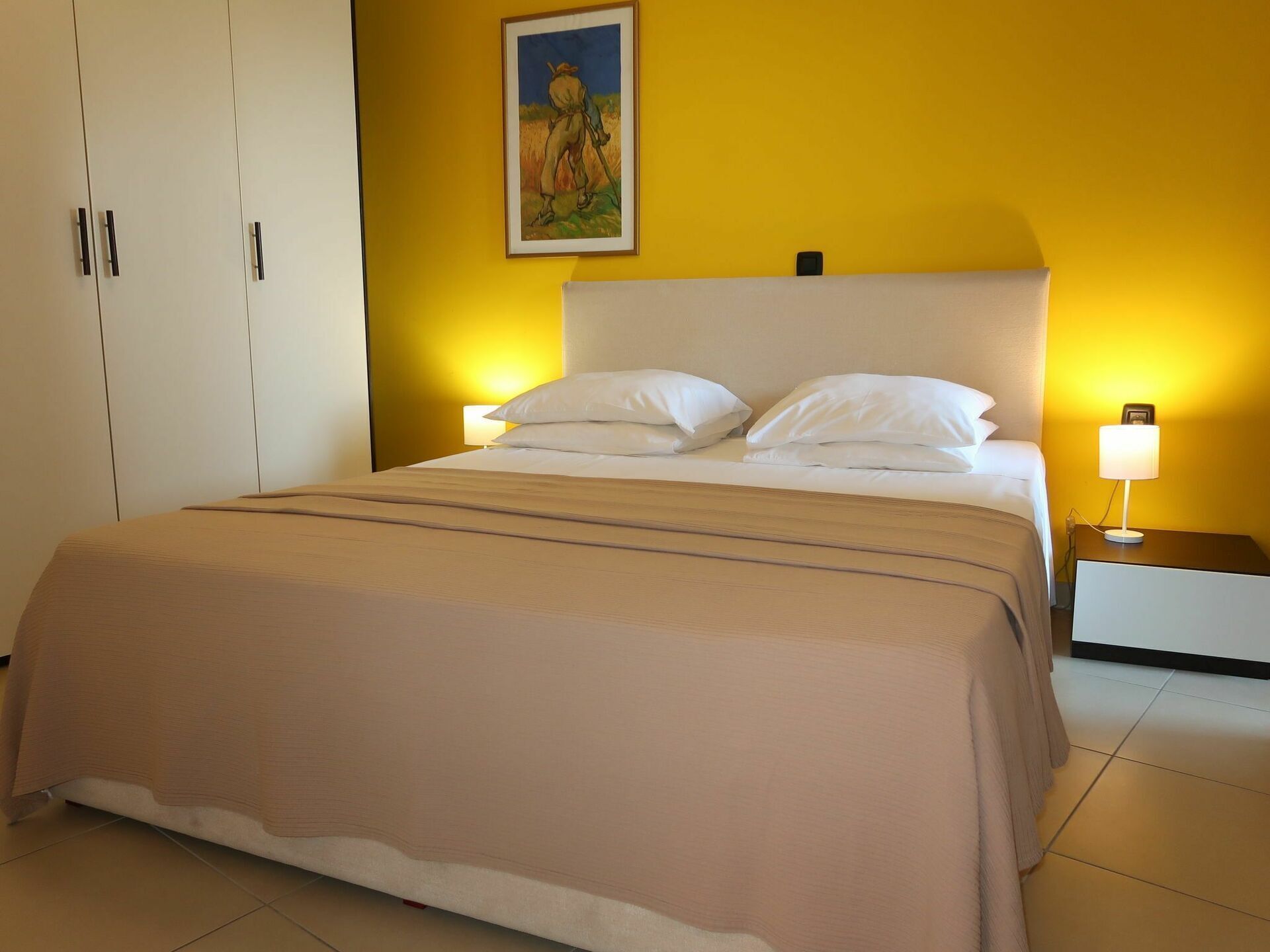 A neatly made bed with a white headboard and two bedside lamps, a painting on the wall, and a wardrobe with a handle.