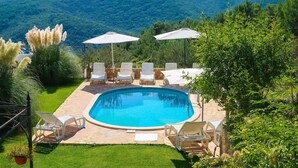 Una piscina con tumbonas y sombrillas, rodeada de un césped cuidado y árboles, con montañas al fondo.